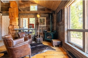 A log cabin interior with a wooden ceiling, a large window, a fireplace, a wooden sculpture, and leather furniture.