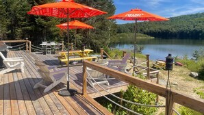 A wooden deck with picnic tables and chairs, surrounded by a lake and forest. - Beautifully Private Chalet in Sainte Anne du Lac! Perfect lake! (Sainte Anne du Lac)
