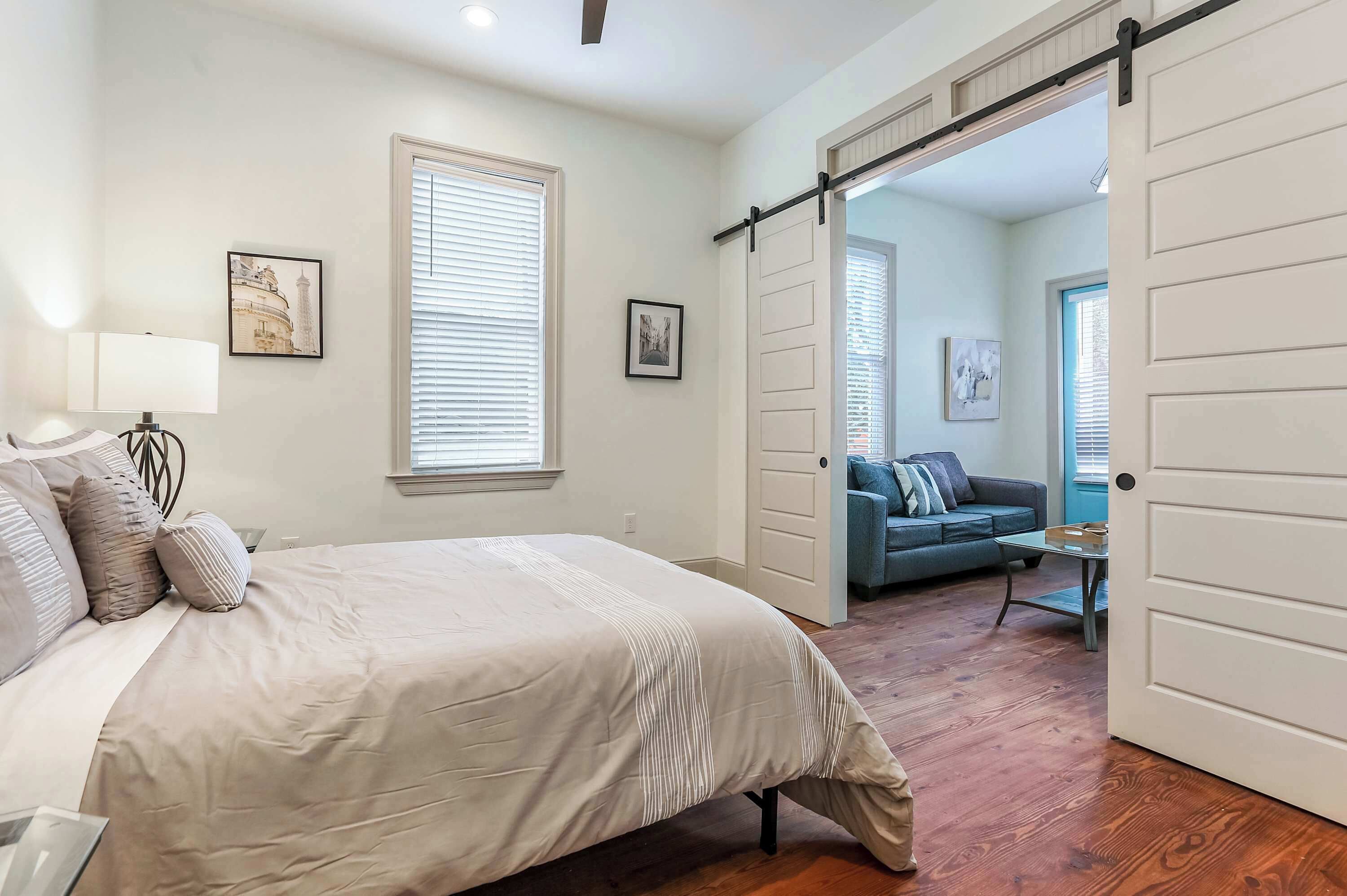 A bedroom with a bed, a nightstand, a lamp, and a window with blinds. There is a door leading to another room.