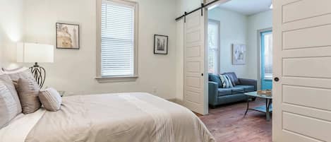 A bedroom with a bed, a nightstand, a lamp, and a window with blinds. There is a door leading to another room.