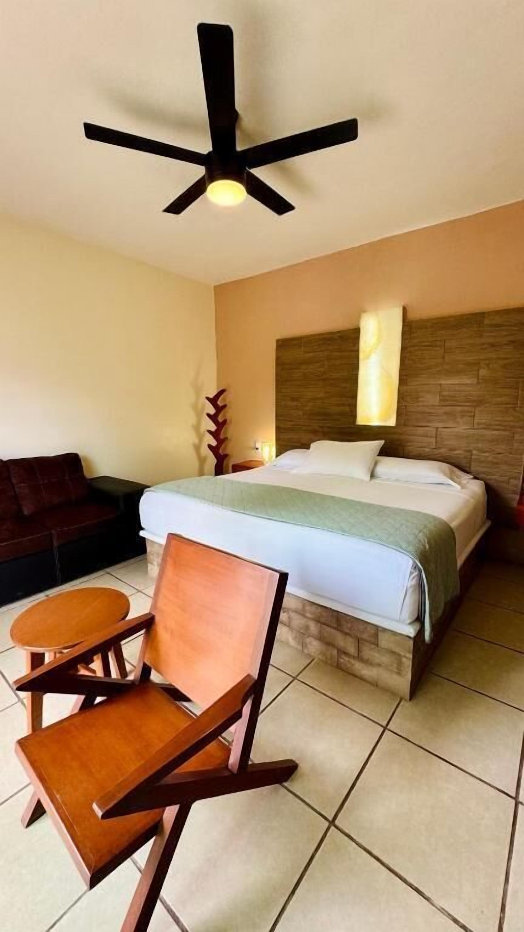 Habitación de hotel con cama, ventilador de techo, una silla de madera, una mesita pequeña y un buró.
