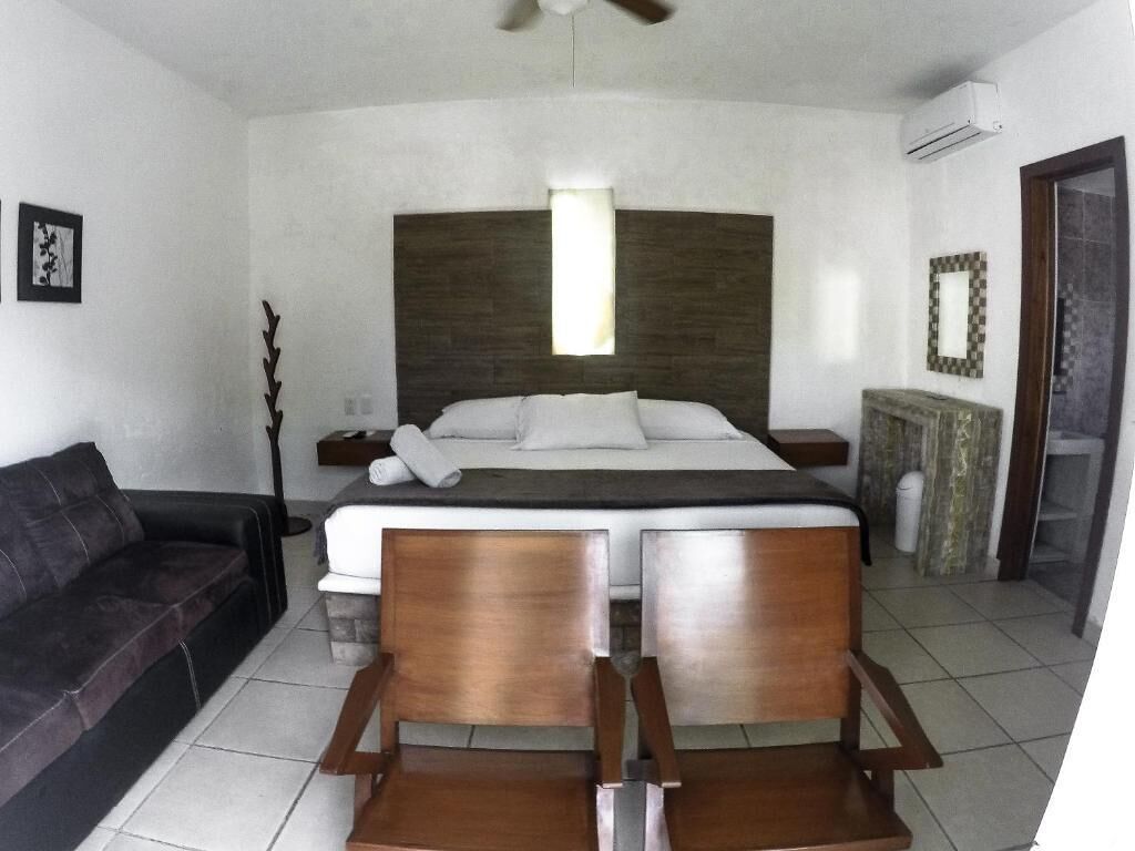 Un dormitorio con una cama, una silla de madera, una mesita de noche, un aire acondicionado instalado en la pared y un cuadro enmarcado.