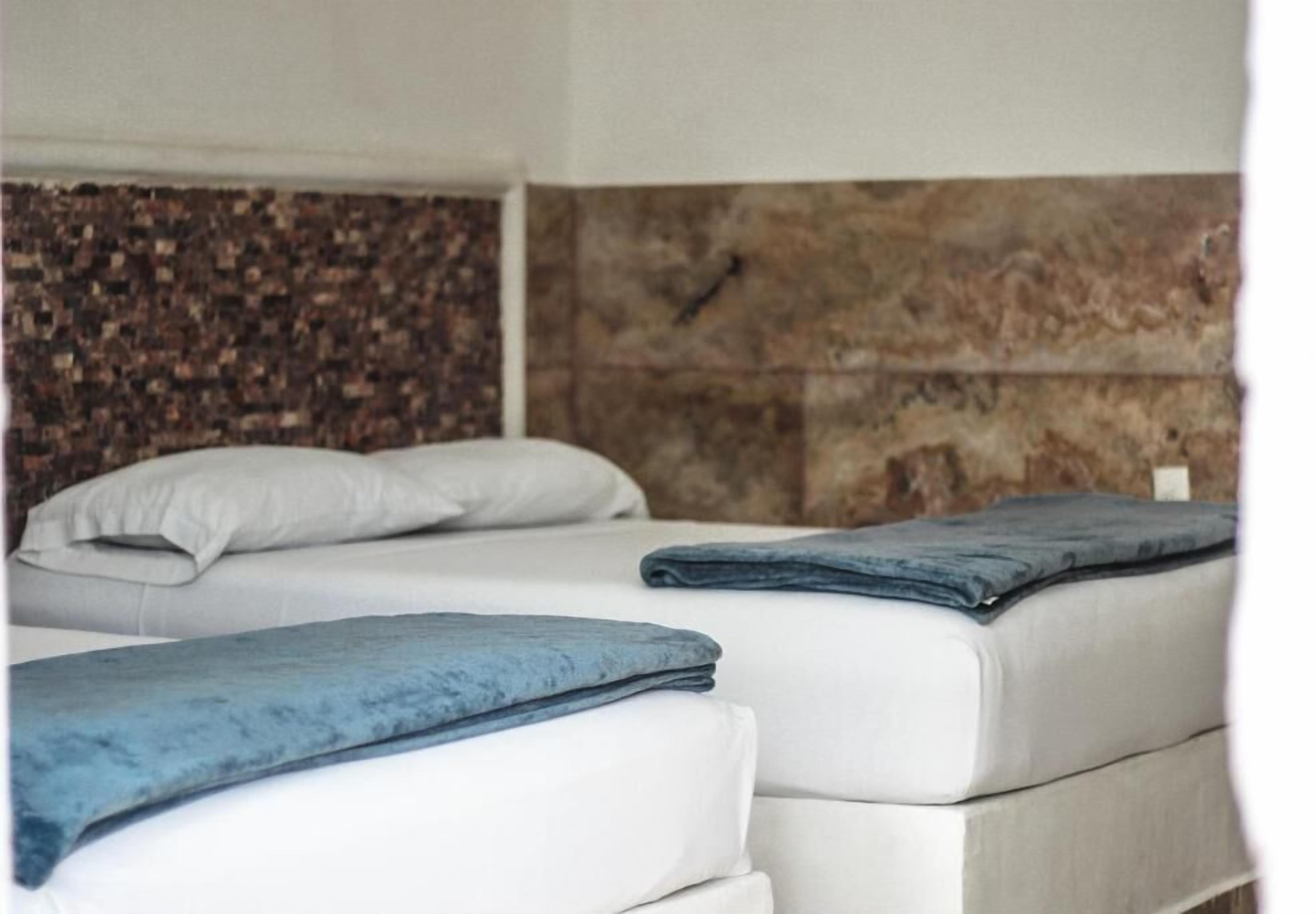 Deux lits avec du linge de lit blanc et des couvertures bleues, un mur texturé en arrière-plan et un petit objet blanc accroché au mur.