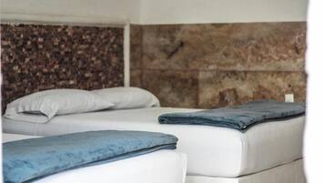 Deux lits avec du linge de lit blanc et des couvertures bleues, un mur texturé en arrière-plan et un petit objet blanc accroché au mur.