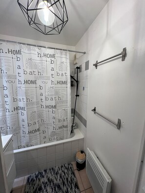 A bathroom with a shower curtain featuring words like 'HOME' and 'HAPPINESS,' a towel rack, a radiator, and a waste bin. - LE NID PROVENÇAL - Ground-floor apartment with garden, pool, and terrace (Le Pontet)