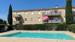 En rektangulär swimmingpool med en stenlagd poolterrass, omgiven av en låg häck och ett staket av metall.