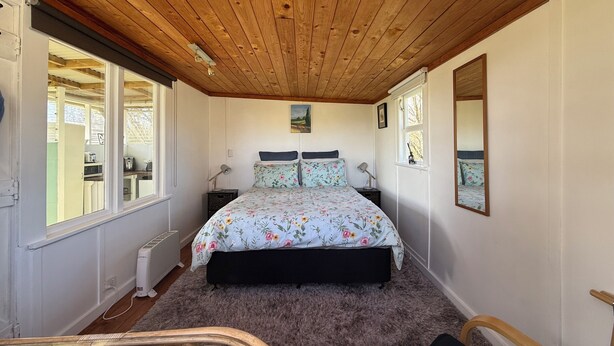 A bedroom with a wooden ceiling, a bed with floral bedding, a bedside table, a lamp, and a window. - Hilltop farm cabin, private deck & sweeping views, 10 mins to Ōhope Beach (Wainui)