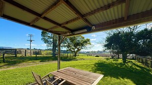 A covered outdoor seating area with a wooden table and chairs, overlooking a grassy landscape with trees and a fence. - Hilltop farm cabin, private deck & sweeping views, 10 mins to Ōhope Beach (Wainui)