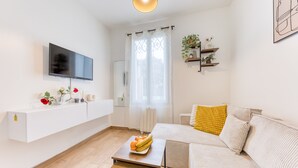 En moderne stue med et fladskærms-tv, en sofa, et sofabord med frugt, et hvidt konsolbord og et vindue med gardiner.