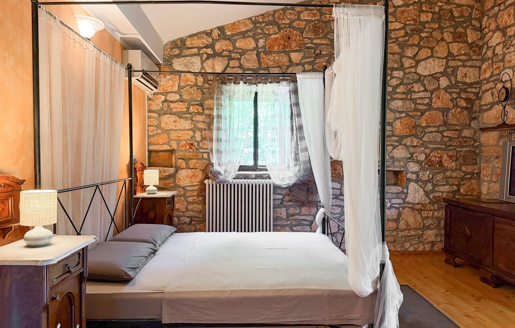 A bedroom with a stone wall, a bed with a canopy, a nightstand, a lamp, and a window with curtains.