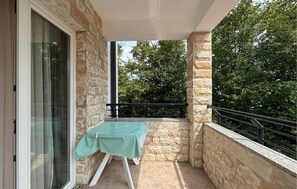 Ein Balkon mit Tisch, der mit einer grünen Decke bedeckt ist, Steinwänden und Blick auf Bäume.