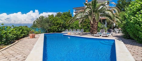 A rectangular swimming pool with a tiled edge, surrounded by lounge chairs and palm trees, with a building in the background.