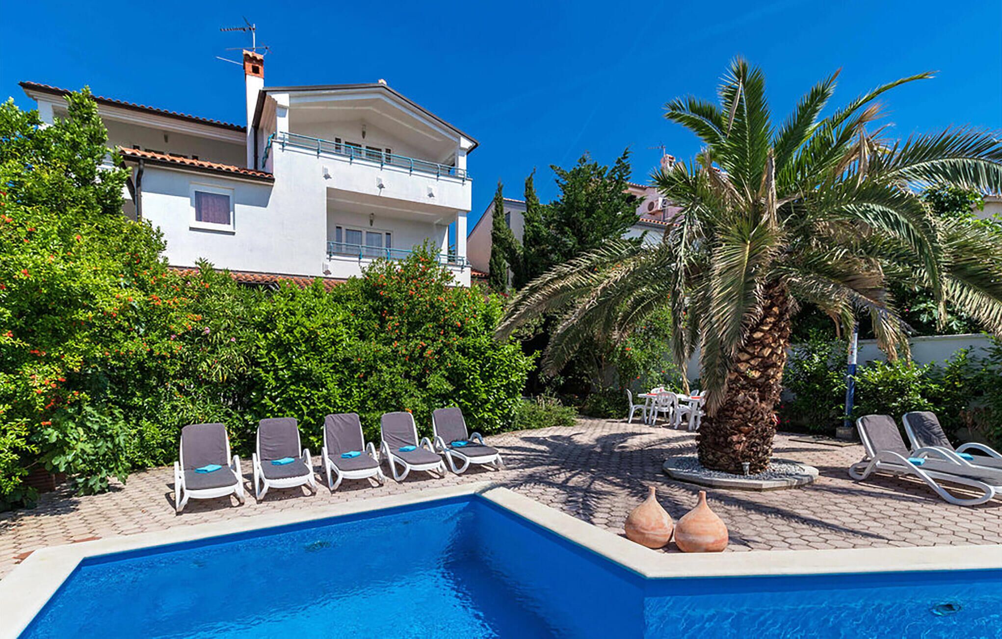 A pool area with lounge chairs, a palm tree, and a building in the background.