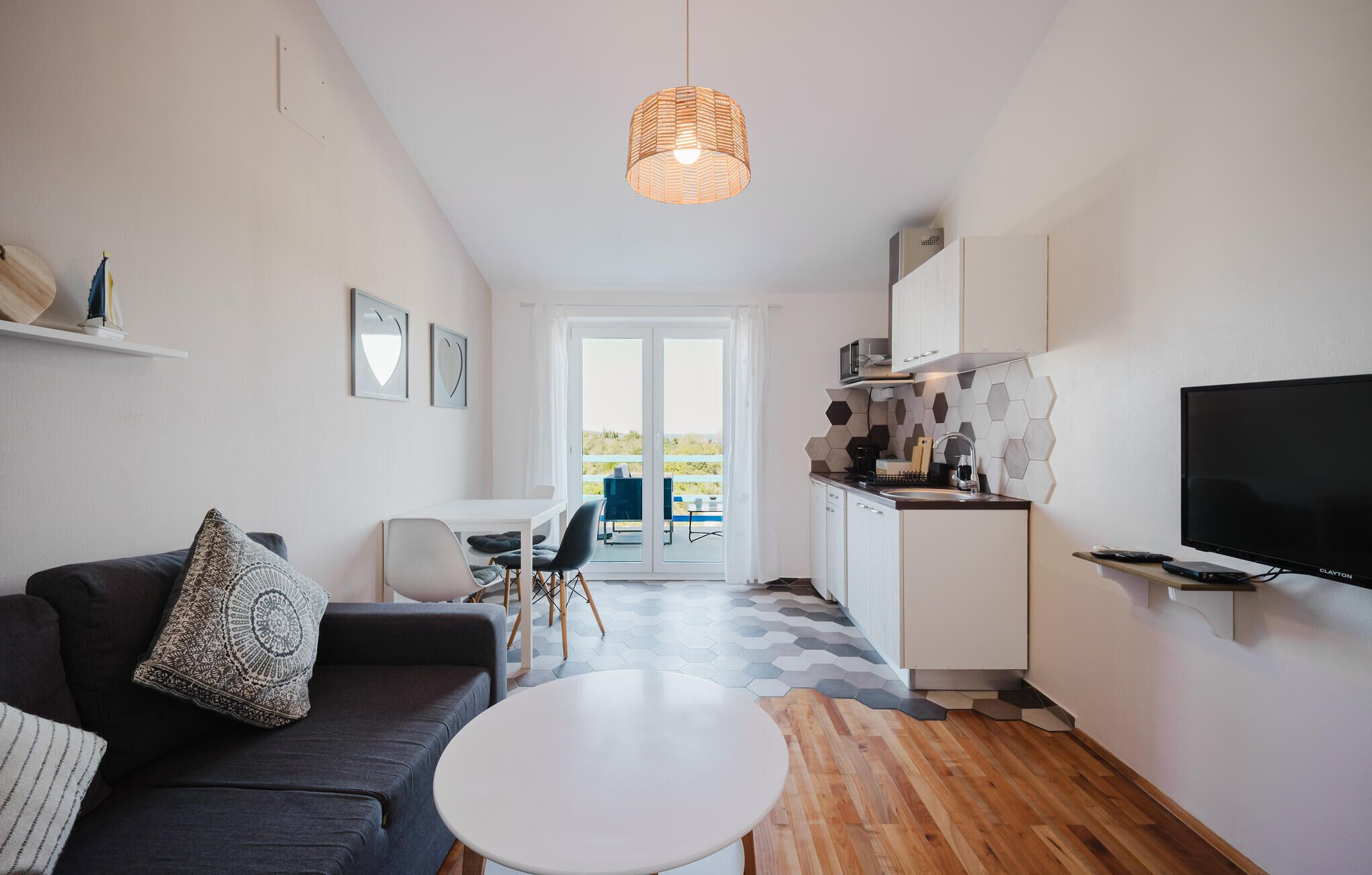 A modern living room with a sofa, a round table, a dining area with chairs, a kitchen with a microwave, and a television.