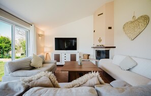 Ein Wohnzimmer mit Fernseher, Kamin und großen Fenstern mit Blick auf einen Garten.