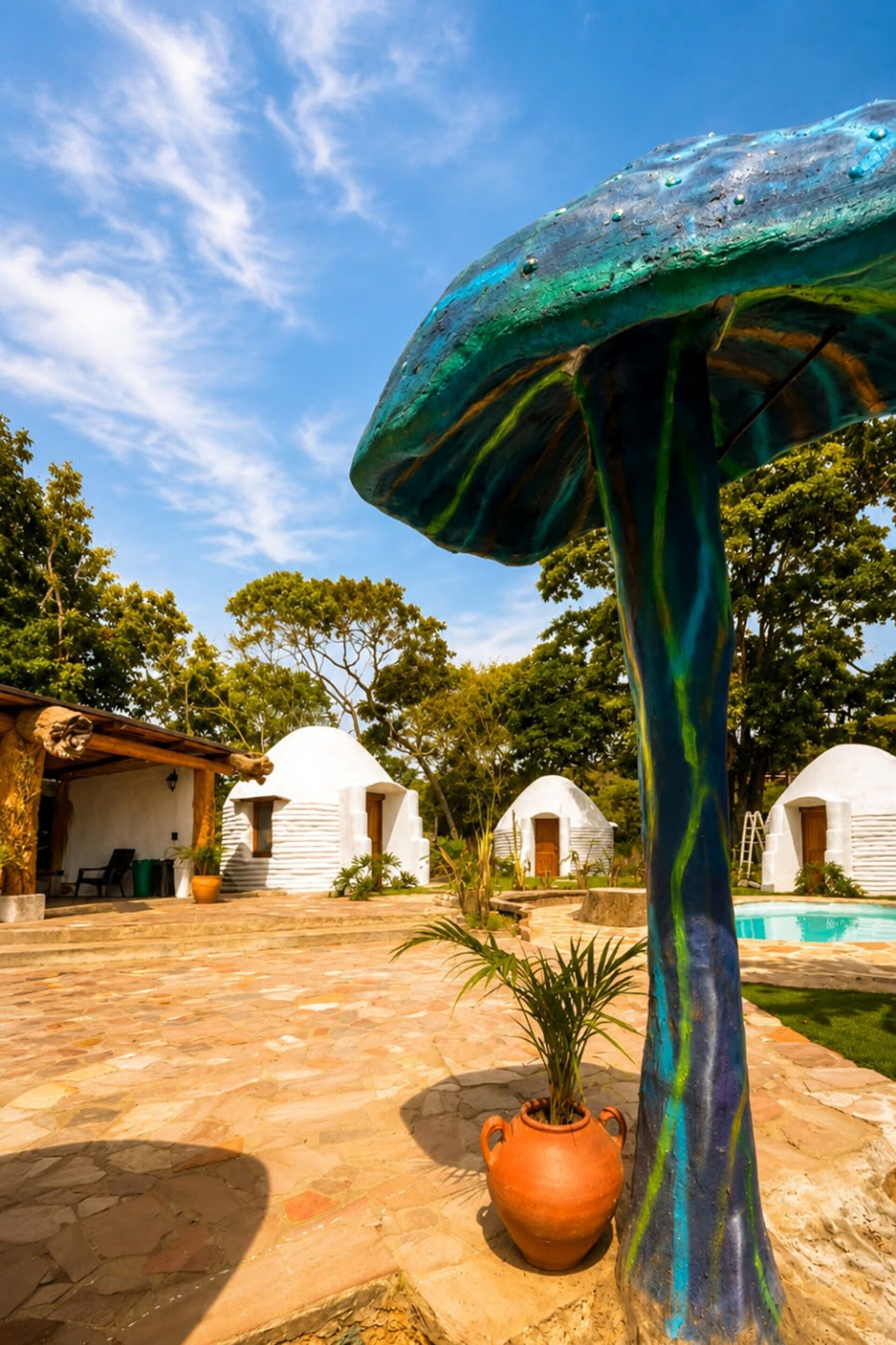 A mushroom-shaped sculpture in a courtyard with white dome-shaped buildings and a pool in the background.