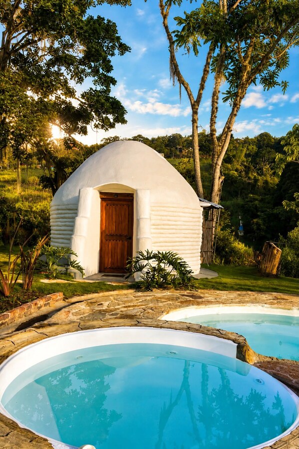 A dome-shaped building with a wooden door, surrounded by lush greenery and two swimming pools.