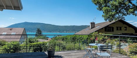 A wooden deck with a table and chairs, overlooking a body of water and mountains.