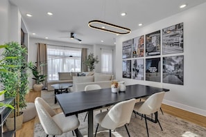 A modern living room with a dark wooden dining table, white chairs, a sofa, and a large window with curtains. - 14 Room, 27 bed, Multi-Townhome Complex, Roof Decks, 2mi to DT! (Nashville)