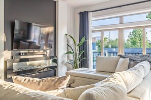 A modern living room with a flat-screen TV, a sofa, a coffee table, and a large window with a view of trees and a building. - 14 Room, 27 bed, Multi-Townhome Complex, Roof Decks, 2mi to DT! (Nashville)