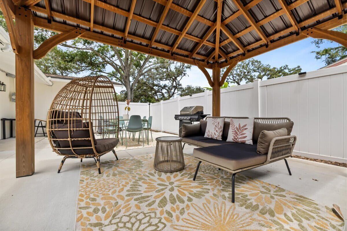 A covered patio area with a wooden pergola, outdoor furniture including a sofa and chairs, and a decorative rug.
