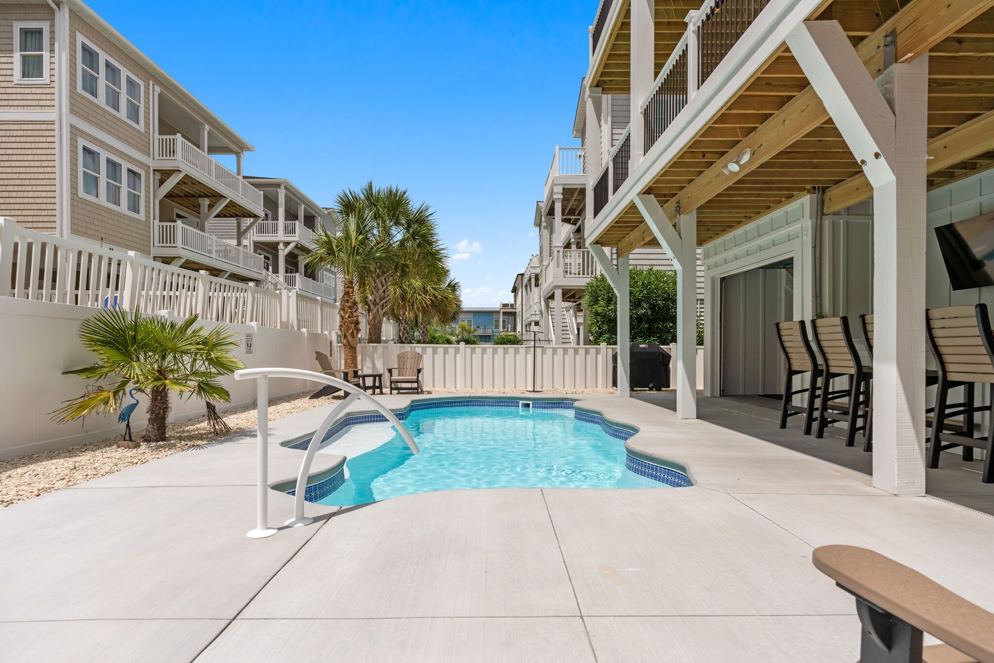 A swimming pool with a wooden deck, a white fence, and a palm tree.
