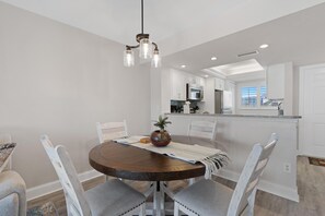 Uma área de refeições com uma mesa redonda, cadeiras brancas, um lustre e vista para a cozinha.