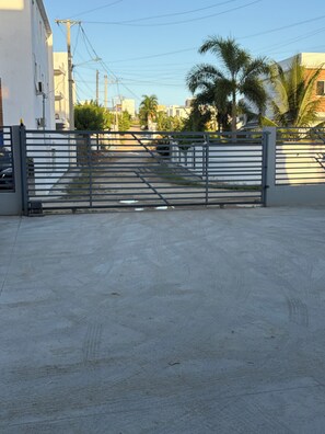 A gated entrance with a metal gate, a concrete ground, and buildings in the background.