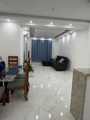 Una sala de estar moderna con comedor, que incluye una mesa de vidrio, sillas de madera, un sofá negro y una cortina azul.