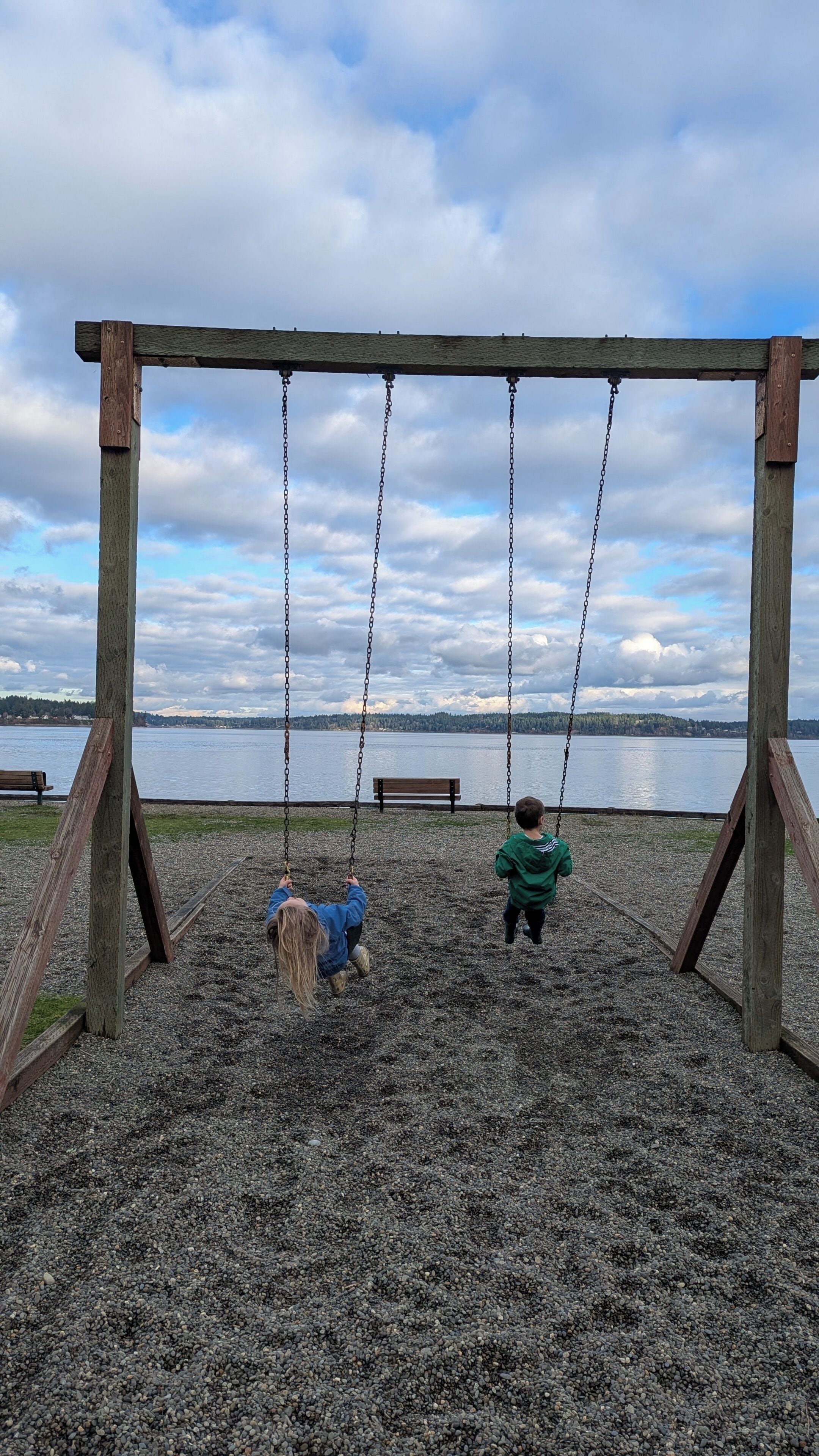 A wooden swing set with a bench, a pebbled ground, and a lake view.