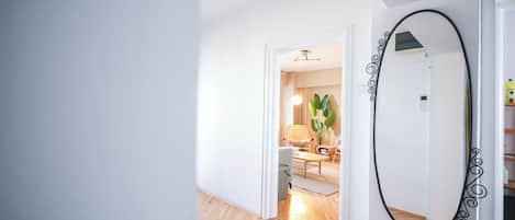 A hallway with a mirror, a door leading to a room with a chair and a plant, and a wooden floor.