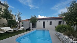 Une piscine avec une terrasse carrelée, un bâtiment blanc doté d’un toit carrelé et du mobilier d’extérieur.