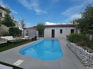 Una piscina con borde de baldosas, un edificio blanco con tejado de cerámica y mobiliario exterior.