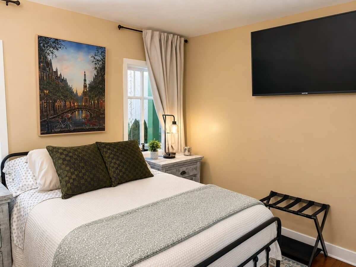 A bedroom with a bed, a nightstand, a lamp, a TV, a window with curtains, and a painting on the wall.