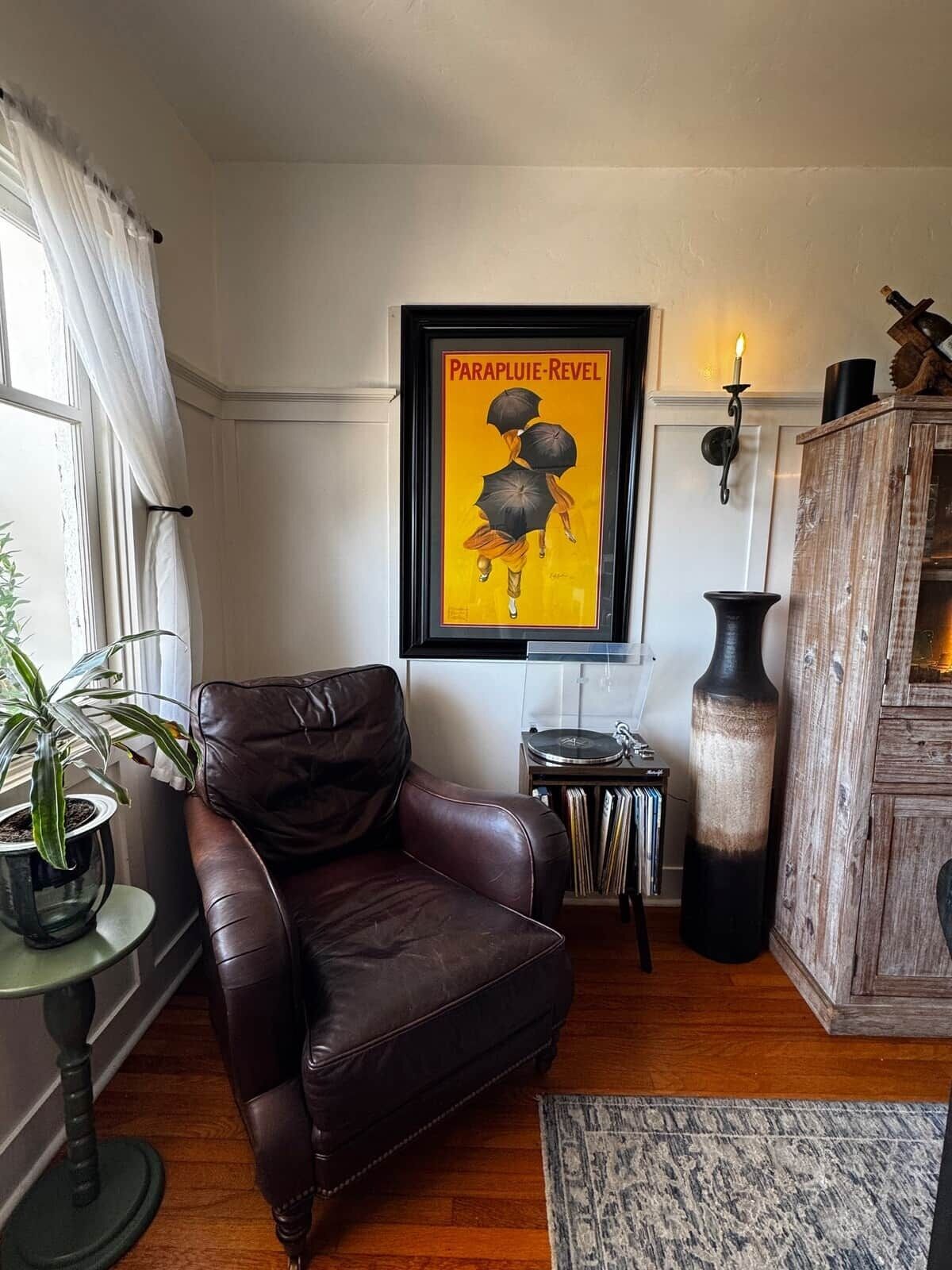 Een woonkamer met een bruine leren fauteuil, een lijst met een poster, een kamerplant, een houten kast en een klein tafeltje met een platenspeler.