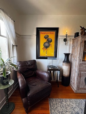 Een woonkamer met een bruine leren fauteuil, een lijst met een poster, een kamerplant, een houten kast en een klein tafeltje met een platenspeler.
