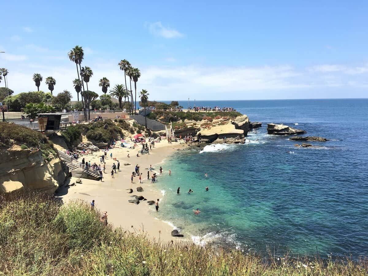 A coastal beach with clear water, palm trees, and rocky outcrops.