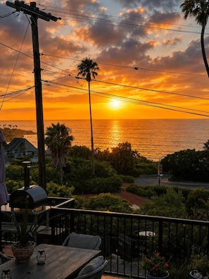 A sunset view from a balcony overlooking a body of water, with a wooden table and chairs, a barbecue grill, and a railing. - Amalfi House/Hot Tub/Pizza Oven/Ocean Views (San Diego)