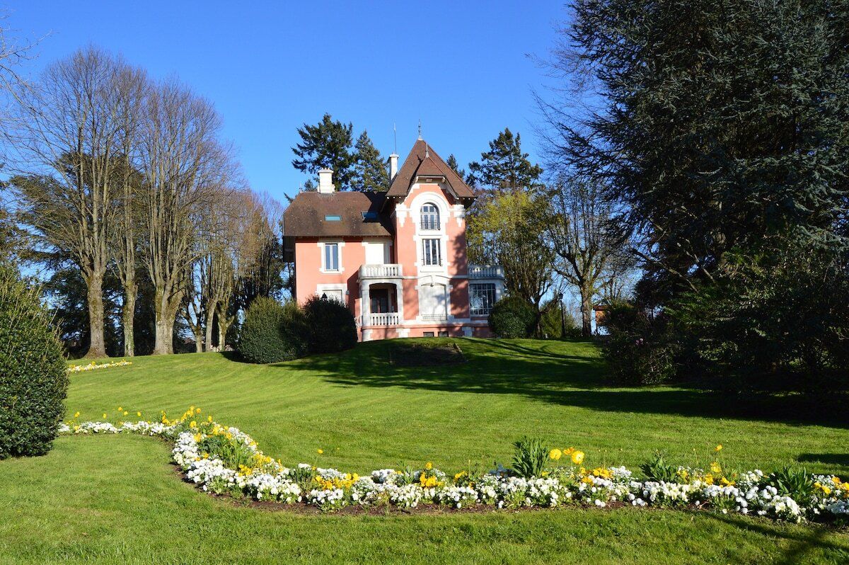Een groot, roze, twee verdiepingen tellend huis met een symmetrische gevel, omgeven door een verzorgd gazon en diverse bloeiende planten.