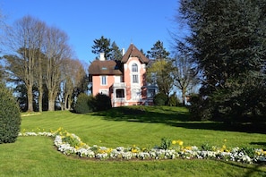 Een groot, roze, twee verdiepingen tellend huis met een symmetrische gevel, omgeven door een verzorgd gazon en diverse bloeiende planten.