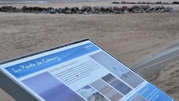 Un cartel informativo en una playa con texto e imágenes, una orilla arenosa y un rompeolas rocoso.