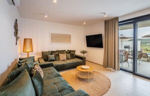 Ein modernes Wohnzimmer mit einer Eckcouch, einem runden Couchtisch, einem Flachbildfernseher und einer großen Schiebeglastür, die zu einem Außenbereich führt.
