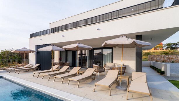 A modern outdoor pool area with lounge chairs and umbrellas, a clear pool, and a building with large windows and a balcony. - Amazing home in Kolan (Kolan)
