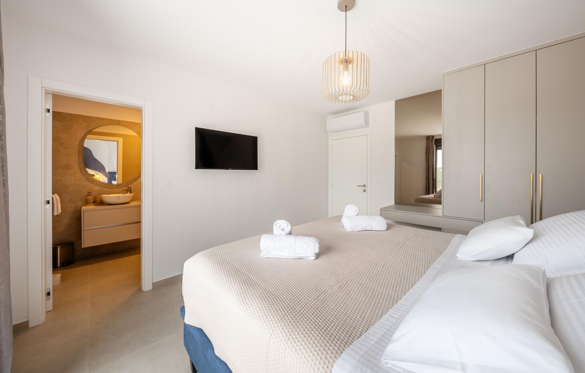 A bedroom with a bed, a TV, a mirror, and a bathroom with a sink and towel rack.
