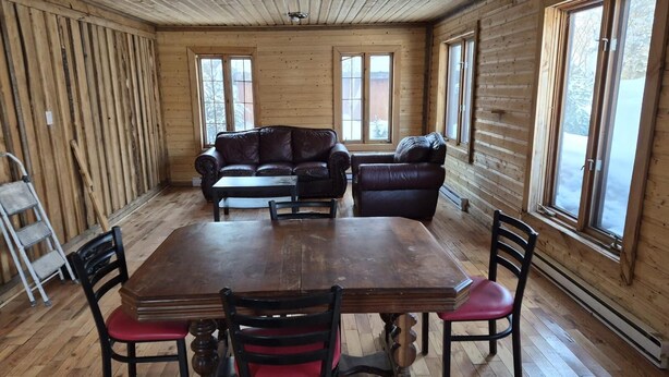 A wooden cabin interior with a dining table, chairs, a sofa, and large windows. - Waterfront Chalet! The Getaway You Need! (Lantier)