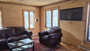 A wooden cabin interior with a leather sofa, a smaller leather armchair, a coffee table, and a large window. - Waterfront Chalet! The Getaway You Need! (Lantier)