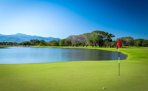 Un parcours de golf avec un mât de drapeau, un plan d’eau et des arbres.