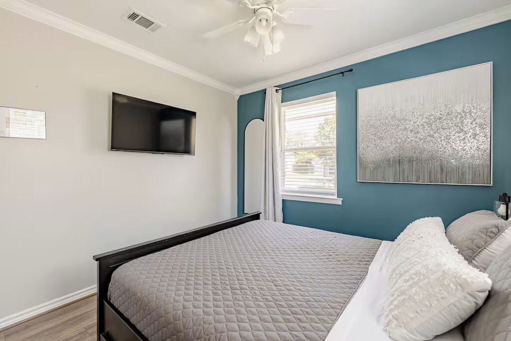 A bedroom with a bed, a ceiling fan, a mounted TV, a window with curtains, and a painting on the wall.