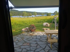 Eine Terrasse mit Steinboden, einem Metallstuhl, einem Holztisch und Blick auf einen See und Wald.
