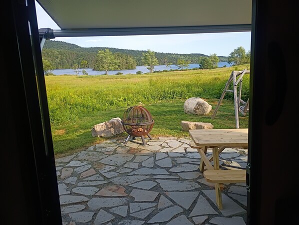 Un patio con piso de piedra, una silla de metal, una mesa de madera y vistas a un lago y bosque.